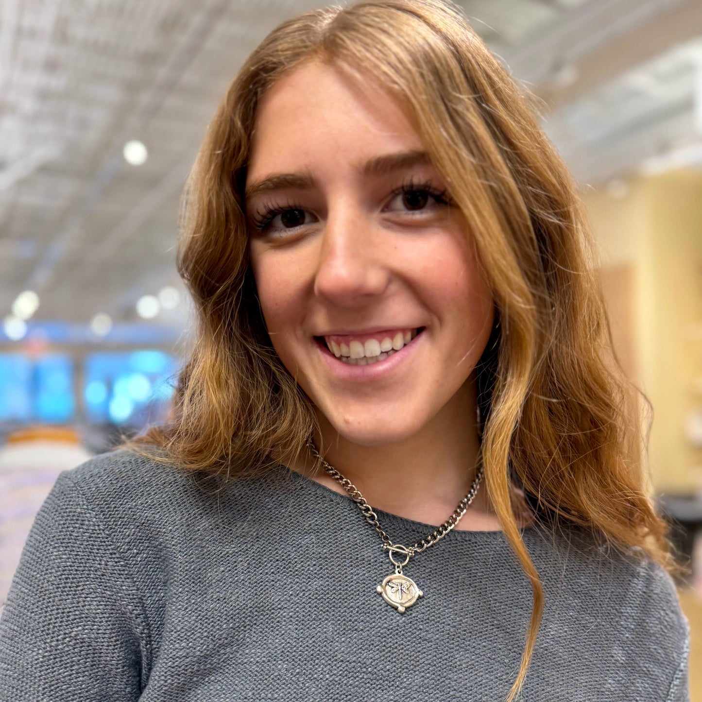 Woman with blonde hair wearing a gray sweater and necklace indoors