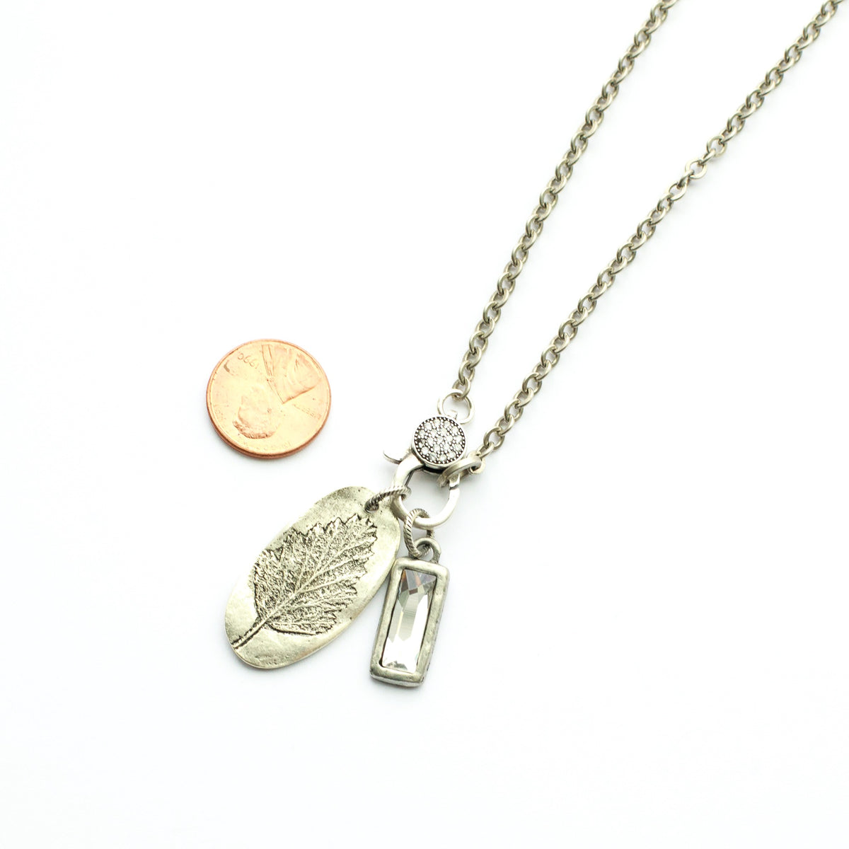 Silver necklace with leaf and rectangular pendant next to a penny on a white background