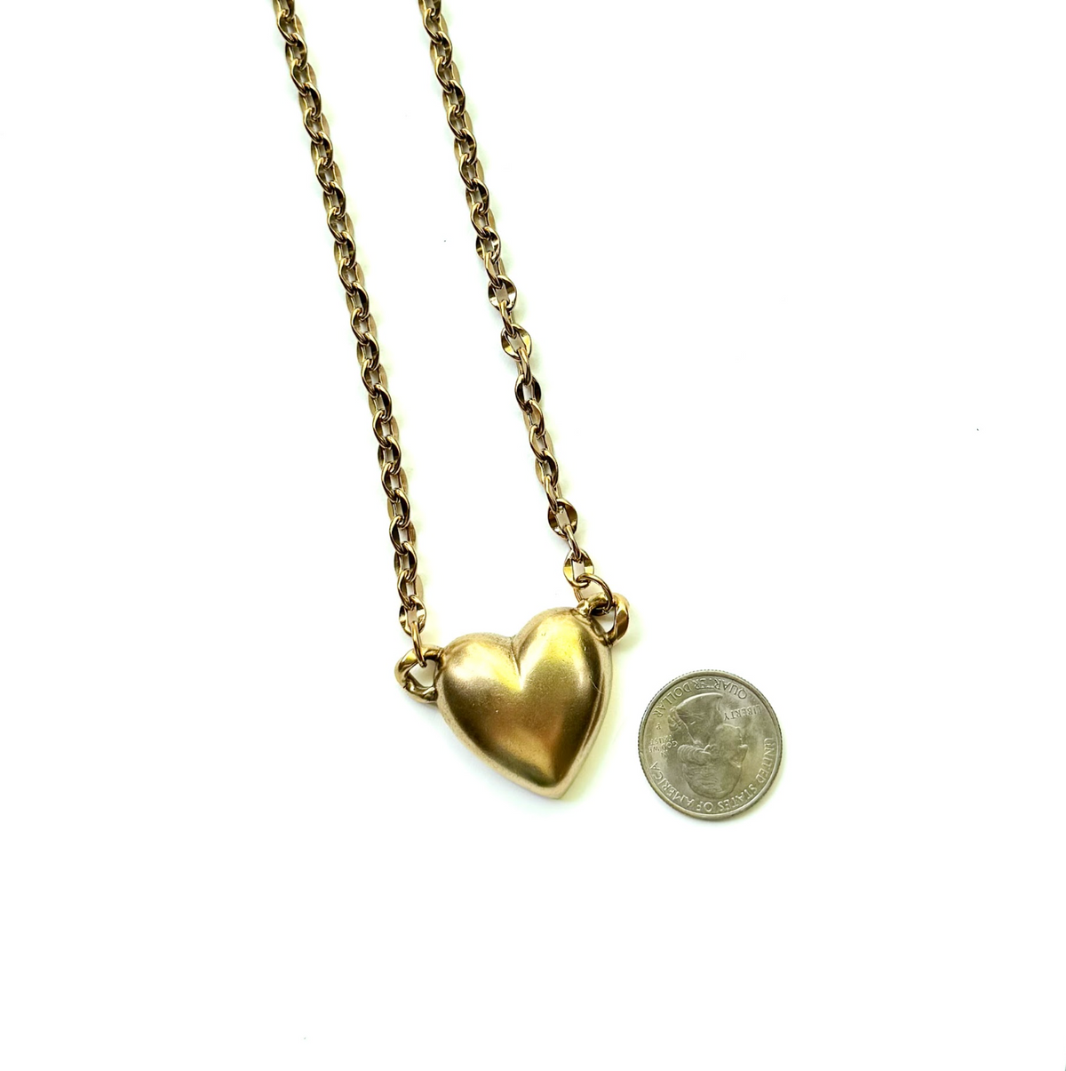 A gold heart pendant necklace on a gold chain, shown next to a US quarter coin for scale on a plain white background.