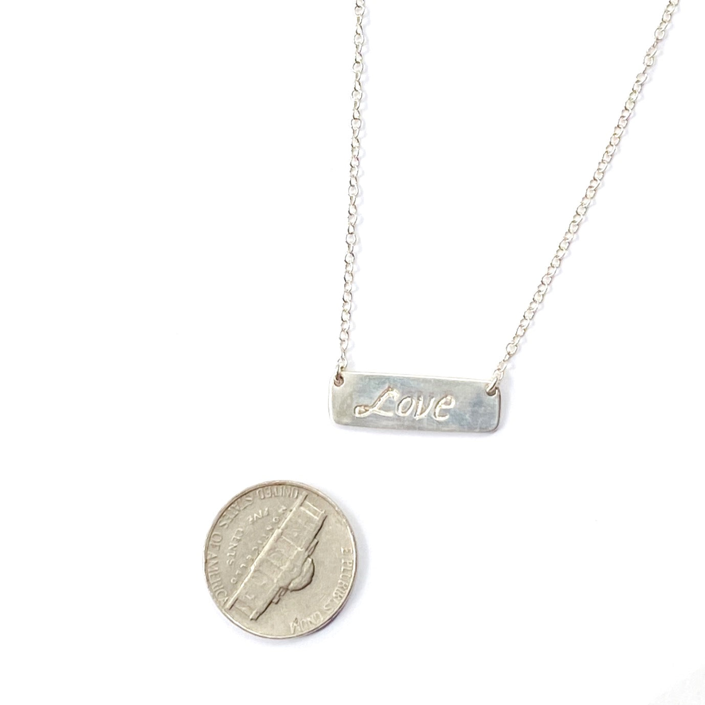 Silver necklace with a bar pendant engraved with 'Love' next to a quarter for scale on a white background