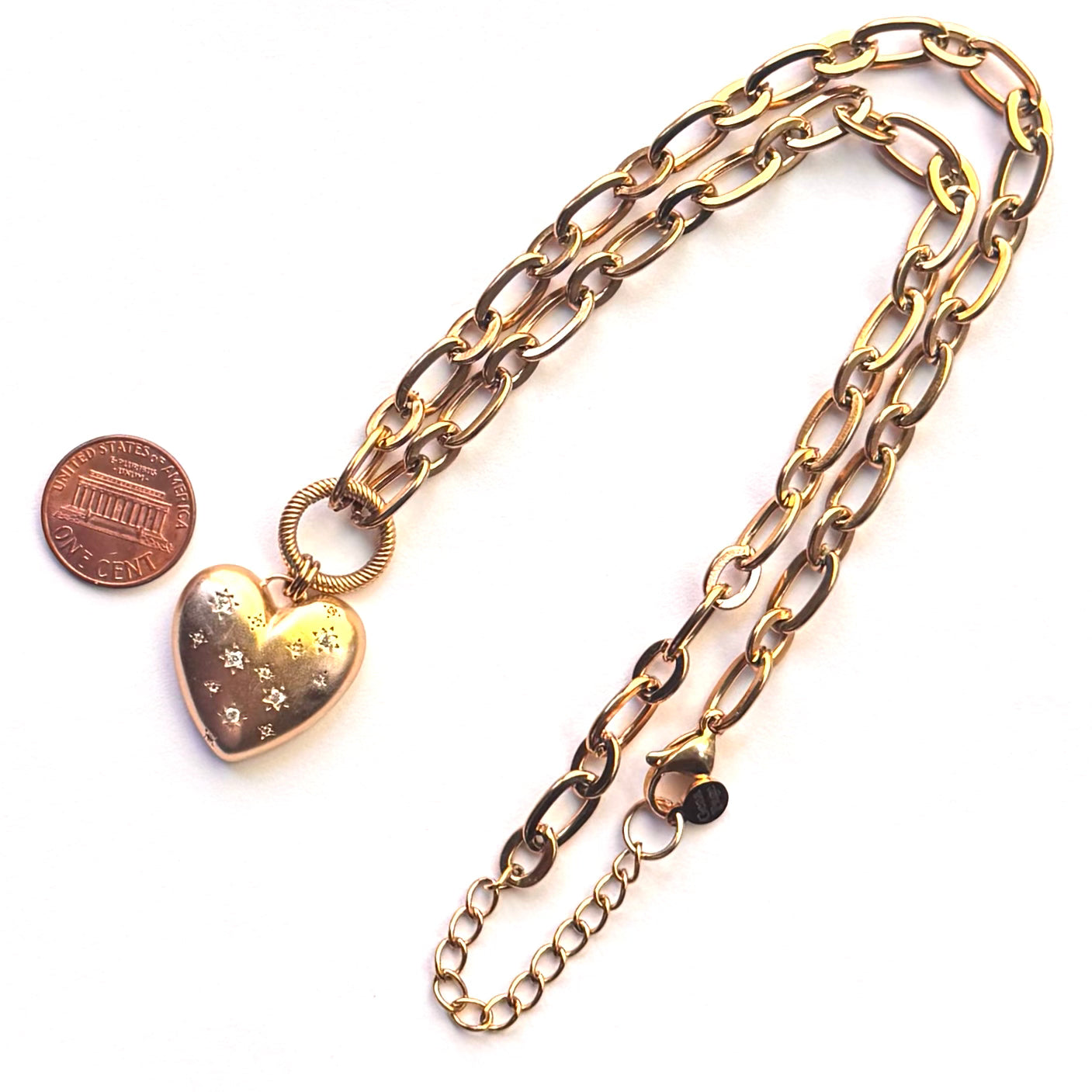 A gold-toned chain necklace with a heart-shaped locket pendant featuring small star engravings, displayed on a white background next to a penny for scale.