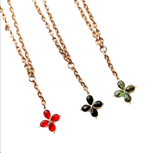 Three gold-tone chain necklaces with four-stone clover pendants are arranged diagonally on a white background. The necklaces feature red, black, and green gemstone pendants.