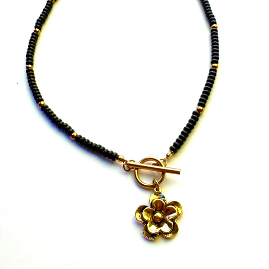 A beaded necklace featuring small black beads accented with gold spacers, a gold toggle clasp, and a dangling gold flower charm, shown on a plain white background.