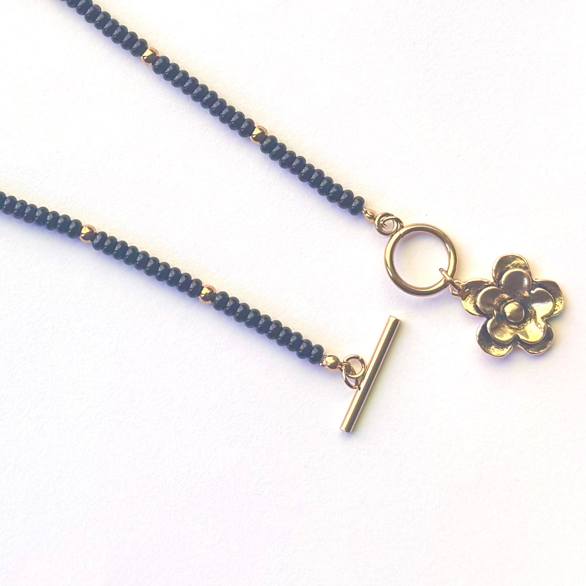 A beaded necklace with black round beads, gold spacer accents, a gold toggle clasp, and a dangling gold flower charm, displayed on a clean white background.