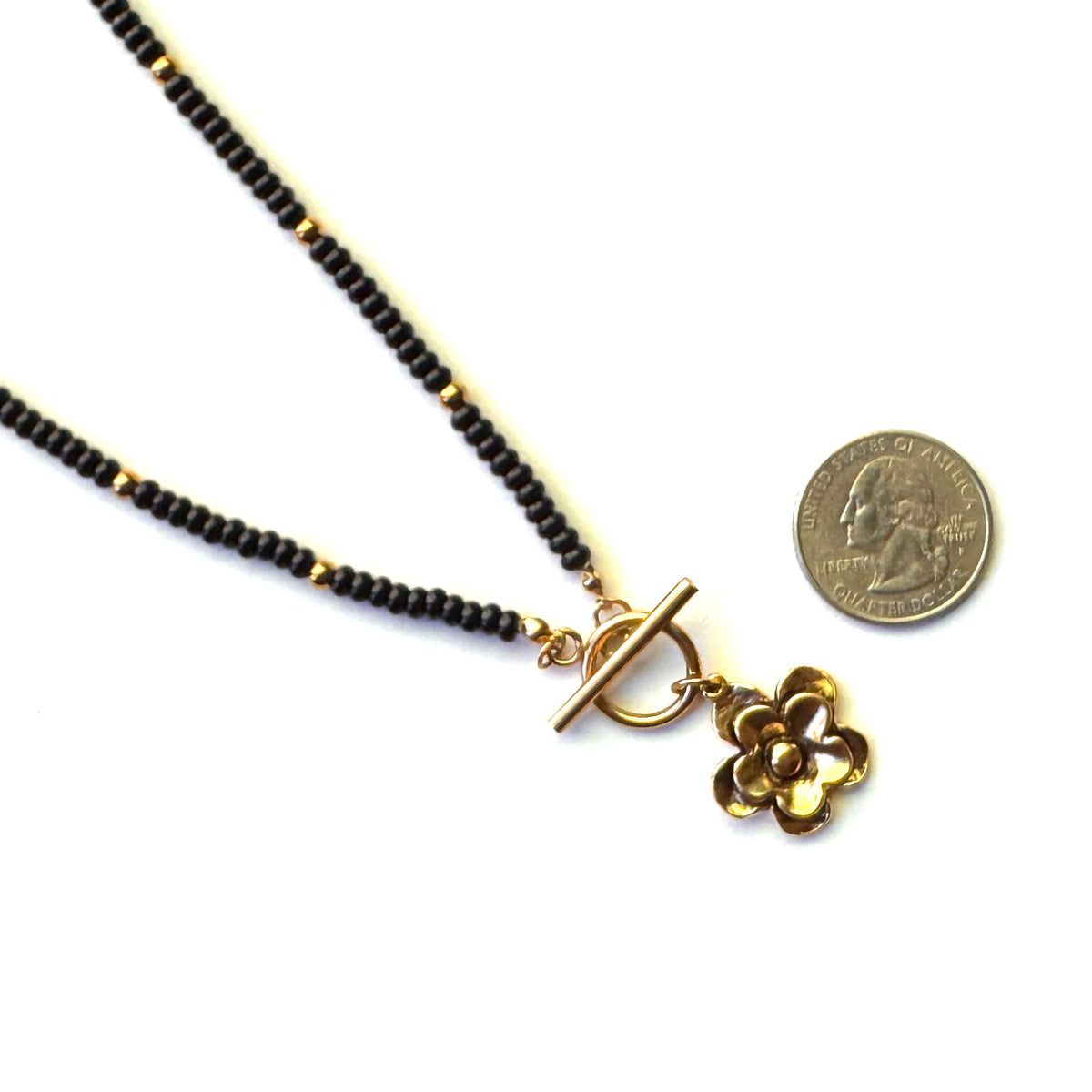 A black beaded necklace with a gold toggle clasp and a small gold flower charm, shown next to a US quarter coin for scale on a white background.