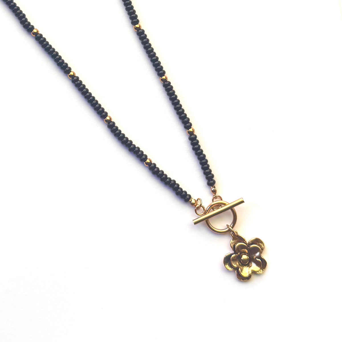A beaded necklace featuring dark gray or black round beads accented with small gold spacers, a gold toggle clasp, and a dangling gold flower charm, shown on a plain white background.