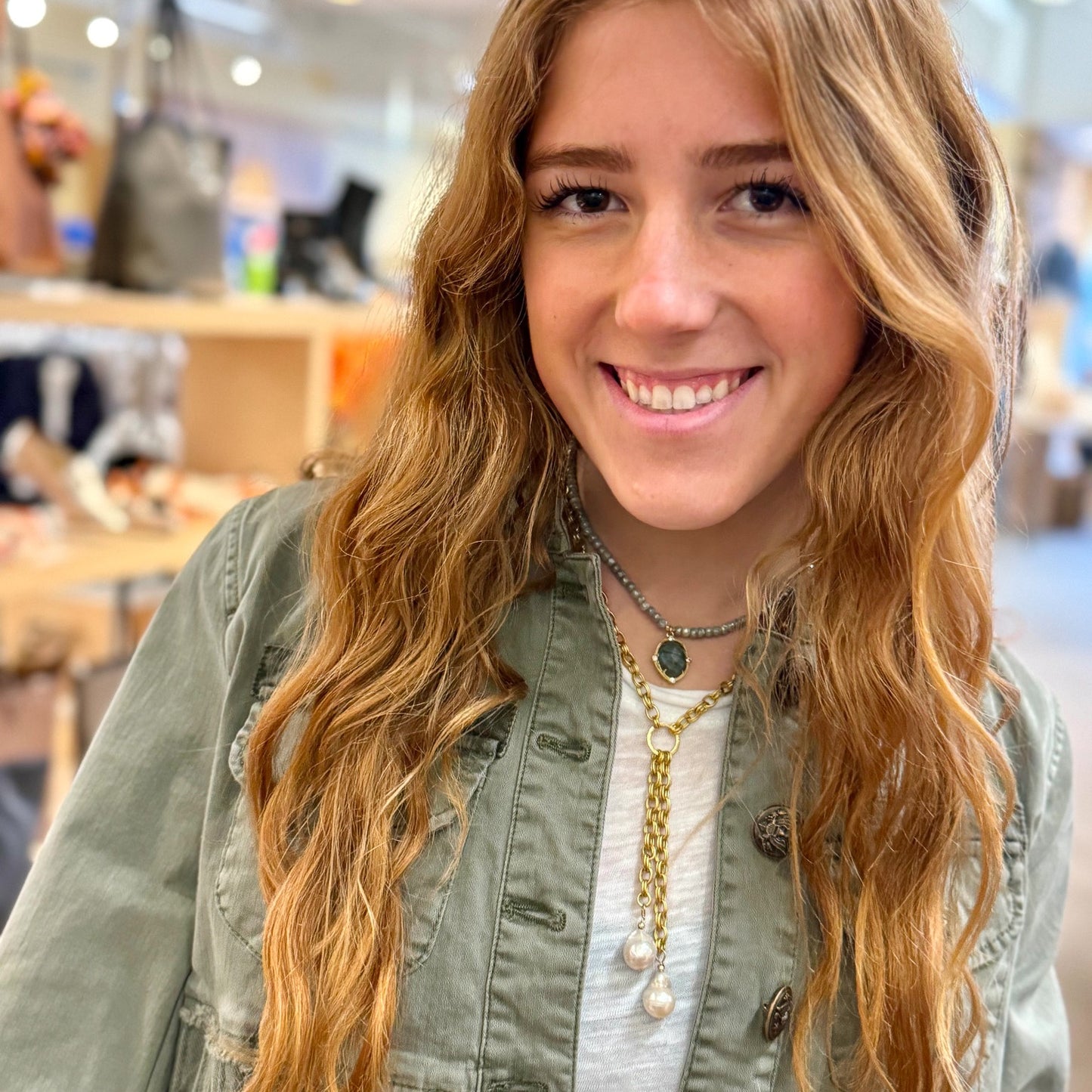 Woman with long blonde hair wearing a green jacket and multiple necklaces in an indoor setting.