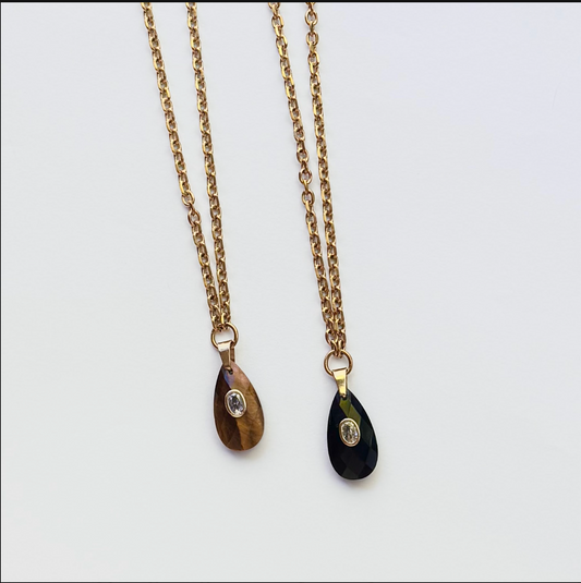 Two gold chain necklaces with teardrop pendants, one featuring a brown cat's eye stone and the other a black onyx stone, both with small clear crystal accents, displayed on a white background.