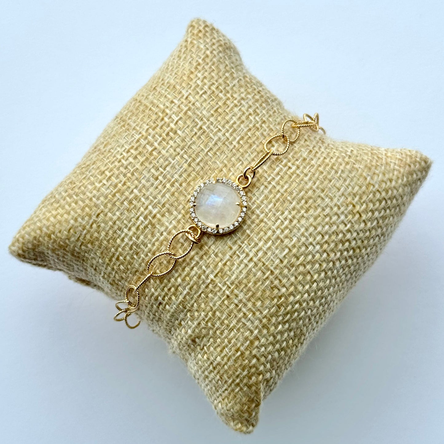 Gold bracelet with a central gemstone on a textured beige pillow against a light blue background