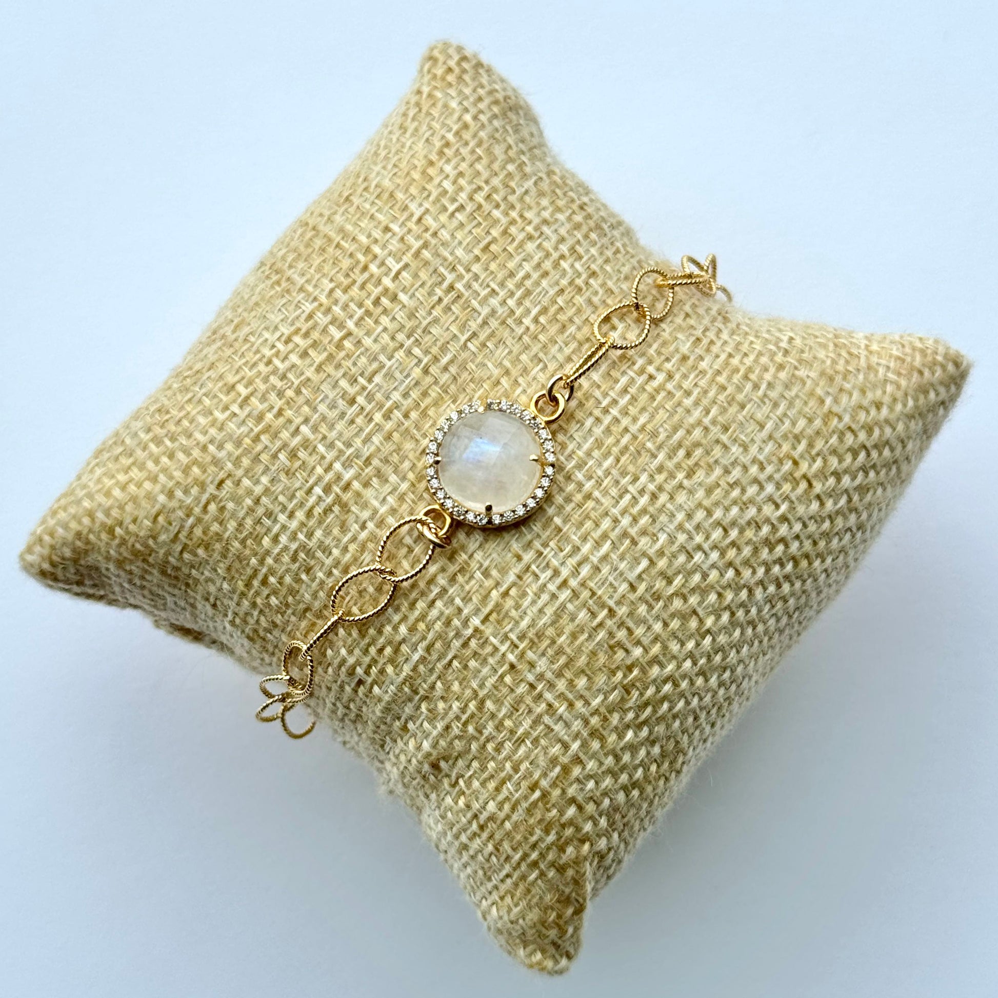Gold bracelet with a central gemstone on a textured beige pillow against a light blue background