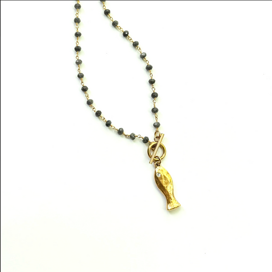 A beaded necklace featuring small dark gray beads on a gold-tone chain, with a gold-tone toggle clasp and a dangling fish-shaped pendant, shown on a plain white background.